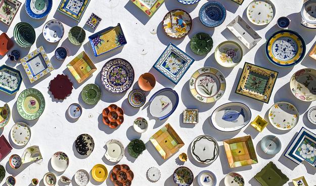 Lots of colour plates and dishes with various designs in bright colours fixed to a white wall on a  Portuguese potters studio. A bright morning sun is casting string shadows at an angle.