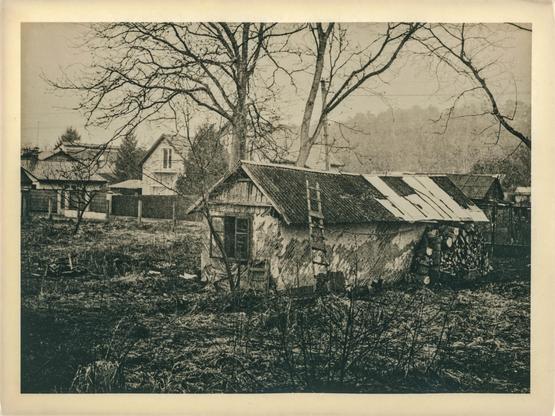 This image is a lith print photograph depicting a rustic rural scene. At the center, there's an old, weathered outbuilding with a corrugated metal roof, some parts of which are patched with lighter panels. A wooden ladder leans against the building, and a neatly stacked pile of firewood sits along its side. Surrounding the structure is an overgrown yard with dry grass and bare shrubs, suggesting early spring.

In the background, several houses with pitched roofs are visible, partially obscured by leafless trees. A forested hillside fades into the mist in the distance, creating a soft, atmospheric effect.

The lith printing technique gives the image its characteristic high contrast, grainy texture, and warm, slightly sepia-toned hues. The shadows are deep and rich, while the highlights are soft and creamy, contributing to the timeless, nostalgic feel of the scene. The broad border around the image also enhances the vintage aesthetic typical of traditional darkroom prints.
