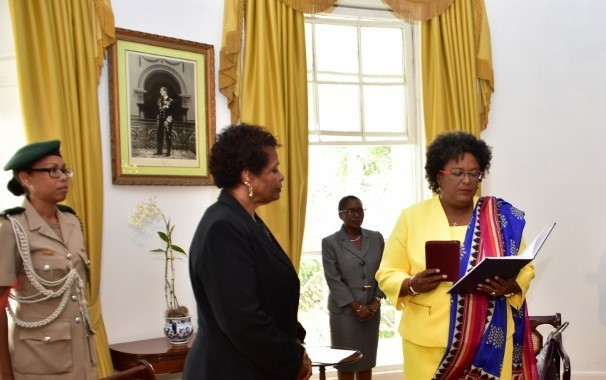 Mia Mottley in 2018 at her swearing in ceremony. She is a black woman in glasses. There are three other women in the state room, all of whom are black and one in military uniform. There is a black and white photo on the wall which might be a former governor or king of England.