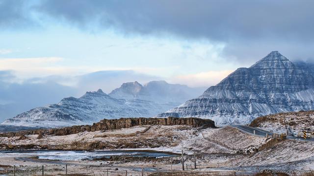 A colour photo of a landscape. The foreground is snowy brown rocks leading down to the waterline on the left. Beyond a rocky ridge with a tarmac road running through it is a beautiful pyramid-shaped mountain, its flanks stepped with eruption layers. Beyond it is another mountain range, craggy peaks capped with snow and ice but with blueish dark rock showing through. The sky is full of snowcloud which is lifting away to the right. Below it is clear cyan sky and banks of cloud above the distant mountains.