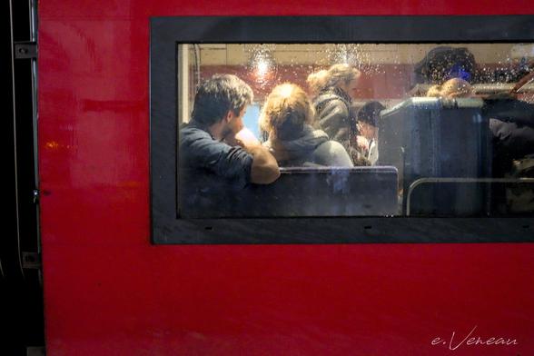 The window of a train, dampened by the rain, reveals the interior: passengers, a woman standing in the middle aisle and a couple sitting with their backs to the window.