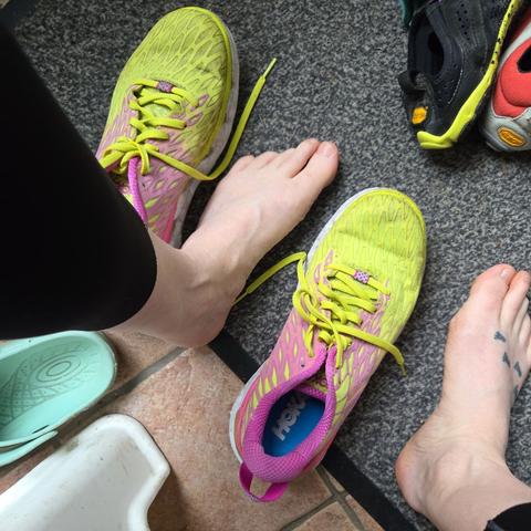 Oppefra foto af et par nøgne fødder der sidder fast på ben klædt i 3/4 løbetights, midt imellem to overvejende gule hokaløbesko, en halv lys turkis wannabe croc, og hælen af to forskellige fivefingers. Det hele befinder sig på fliser og en måtte, med et lysindfald der antyder en dør i nærheden.