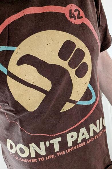 Ein T-Shirt, mal nicht schwarz, sondern braun. Darauf das #HitchhikersGuide Logo (eine Hand zeigt das bekannte Anhalter-Symbol, dahinter ein Planet mit einer Orbitalline drumherum). Das Logo ist mit einem großen roten Kreis umrundet und trägt in der rechten oberen Ecke die Nummer 42.

Unter dem Logo steht in großen Lettern:

DON'T PANIC

Darunter in kleinerer Schrift. 
42 THE ANSWER TO LIFE, THE UNIVERSE AND EVERYTHING