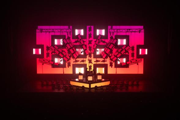 Justice's live setup, a big light rig is illuminated in a pink-red-yellow gradient, reminiscent of a sunset, squares in front of the light rig are illuminated in white. Xavier and Gaspard are comparatively tiny, standing in front.