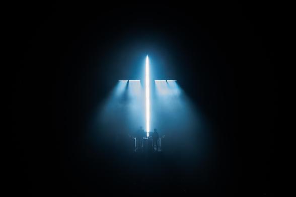Justice's iconic cross made of light bars shining a cool light blue color. Xavier and Gaspard are standing at the bottom of the cross.