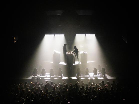 Xavier and Gaspard of Justice, in the darkness, illuminated by two "lamp bars" shining on them, the audience in the pit is visible as well.
