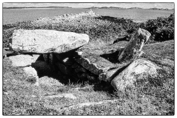 The entrance to a bronze age burial chamber with large stones and a capstone above. In the background is the sea and several islands on the horizon.