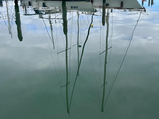A crystal-clear reflection of a two-masted boat in perfectly still water. The white hull of the boat runs along the top of the frame, and the black masts, without sails but with lots of lines of rigging at various angles, are pointing downwards and their tops almost touch the bottom of the frame. The cloudy sky above is rendered various shades of pastel bottle-green in the lagoon water. The image feels like a glimpse into a literal upside-down world, which frankly looks a lot more peaceful than our own.