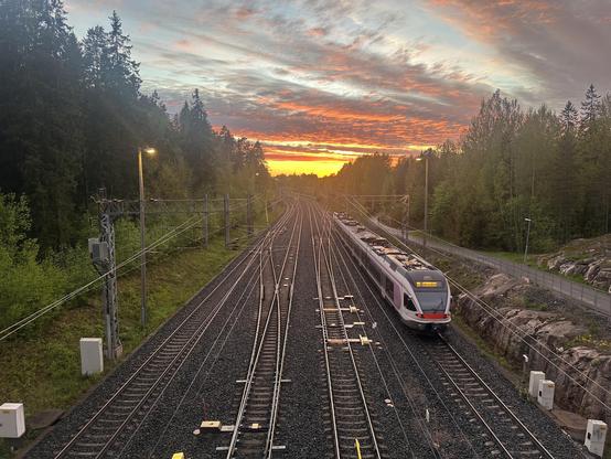 Sm5 train on the rightmost track of a quad-track segment. Above is a pretty sunset with a yellow part mixed with blueish light grey clouds. A bike parh and some greenery is shown on the side.
