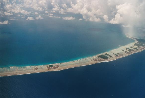 Vue de l'atoll de Rangiroa dans l'archipel des Tuamotu depuis l'avion, photo prise en 2001. On voit une longue bande de terre, qui est en fait du corail affleurant qui s'est couvert de terre et d'un peu de végétation et qui marque la limite entre le lagon, peu profond et bleu clair, en haut sur la photo, et la mer profonde et bleu foncé, en bas sur la photo. L'ensemble forme un grand anneau étroit partiellement immergé, dont on ne distingue qu'une partie sur la photo.