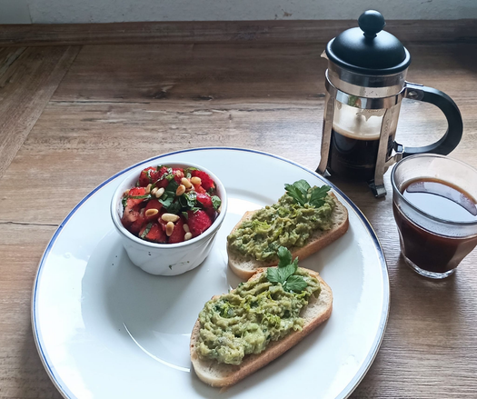 ein Teller mit 2 Scheiben Brot mit Guacamole
eine kleine Schale mit den zubereiteten Erdbeeren. 
Eine kleine French-Presso-Kaffeekanne mit Kaffee und ein Kaffeeglas mit Kaffee