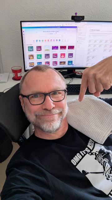 Ein Mann sitzt vor einem Computerbildschirm und zeigt mit dem Finger auf seine Schulter, auf der ein weißes Handtuch liegt. Der Bildschirm zeigt verschiedene Designvorlagen mit der Frage ‚What will you design today?‘. Er trägt ein schwarzes T-Shirt mit einem weißen Aufdruck von R2D2 und dem Text ‚OMG IT'S R2D2‘. Auf dem Schreibtisch sind eine Tastatur, eine Maus, ein roter Becher und ein Notizbuch zu sehen.“