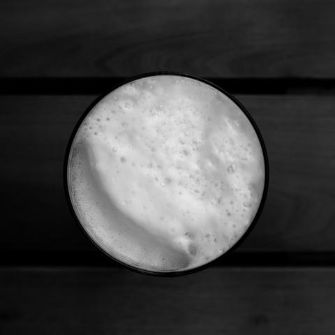 Gefülltes Bierglas mit Schaum von oben fotografiert. Das Foto ist schwarzweiß. Das Glas steht auf einem Biertisch und ist im quadratischen Format.
