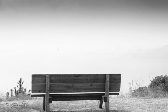 This Is An Abstract / Landscape Black And White Photo In Landscape Format. In The Foreground, Just A Few Metres Away And At Right Angles To The Camera: Stretching Along The Base Of The Image And Offset Very Slightly To The Left Of Centre Is An Empty Wooden Park Bench. There Is Short Grass Surrounding The Bench With Some Small Shrubs To Either Side. Beyond, The Ground Falls Away Rapidly Though This Is Hard To See Since There Is A Large Bank Of Sea Mist Restricting The Visibility. Along the Right Margin And Half Way Up Is A Hint Of Terrain That Slopes Sharply A Short Way Towards The Centre Of The Frame. Either Side Of Centre And A Few km Away Is The Partially Visible View Of Two Bridge Support Towers With A Short Length Of Deck Below. Above The Sky Is Obscured The Light Darkening Slightly Towards The Top Of The Image.
The Location: Angel Island, California. The Bridge: The Golden Gate (2016).