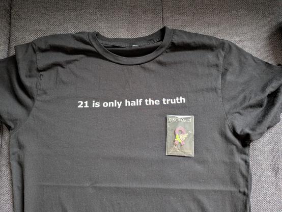 T-Shirt mit der Aufschrift "21 is only half the truth" darauf liegt eine Anstecknadel in Violett mit einem goldenen Flieder