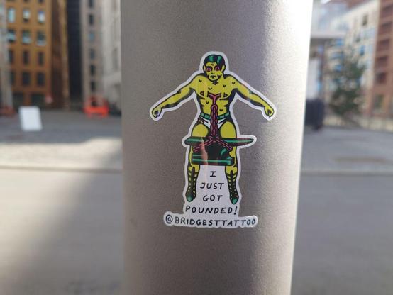 A sticker featuring a yellow cartoon character with a chain-bound torso attached to a weight, standing with arms outstretched. The text below reads 'I JUST GOT POUNDED!' along with '@BRIDGESTTATTOO.'