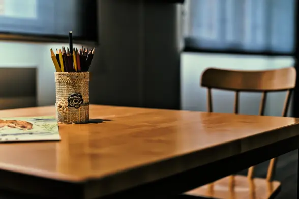 Auf einem Tisch steht ein Becher, der mit Zeitungspapier umhüllt ist, darauf zwei gehäkelte Blüten und ein gehäkeltes Band am oberen Rand. In dem Becher stecken zahlreiche Buntstifte. Links vom Becher ein bemaltes Stück Papier. Rechts am Tisch steht ein Küchenstuhl aus Holz. Das ganz steht in einem Museum und soll Kindern Beschäftigung bieten.