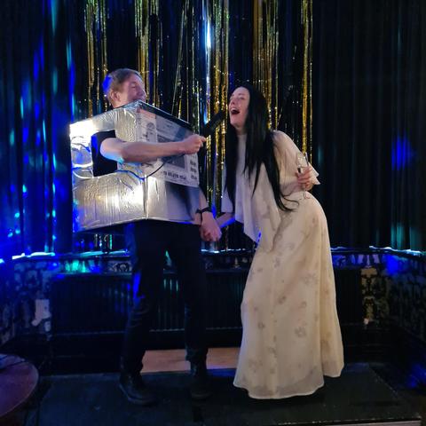 Man in a foil covered cardboard box holding a microphone, into which a woman with a long black wig and 1970s gown is singing.