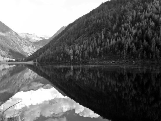 Eine nach Schwarzweiß konvertierte Aufnahme. Die obere Hälfte des Fotos dominiert der Fuß einer nach rechts ansteigenden bewaldeten Erhebung. Jene spiegelt sich in einem die untere Bildhälfte einnehmenden See. Hinter dem See ist links der Erhebung ein Taleinschnitt, der den Blick auf in der Ferne liegende weitere Berge freigibt. Der Himmel ist überbelichtet, in der Spiegelung im See sind aber Wolken und blauer Himmel zu erkennen.