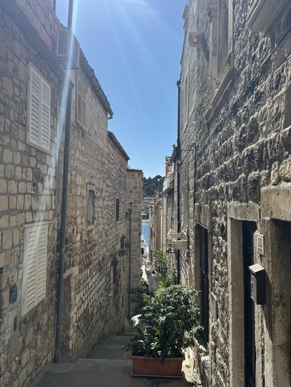 A narrow stone alleyway in a historic Dalmatian town, Hvar in Croatia, lined with old stone buildings featuring shuttered windows and air conditioning units. A potted plant with lush green leaves sits in the foreground on a descending stone staircase. Sunlight beams through the clear blue sky, casting a bright flare across the upper left of the image. At the end of the alley, a glimpse of the sparkling blue sea and distant greenery is visible.