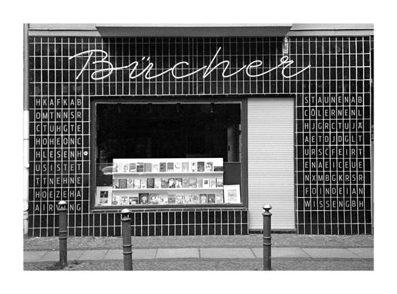 The black and white photo shows the front of a bookshop. "Books" is written in curved lettering above the shop window. The shop window is filled with books. On the sides of the shop window are tiles with randomly arranged letters of the German alphabet. There are two bollards in front of the store.