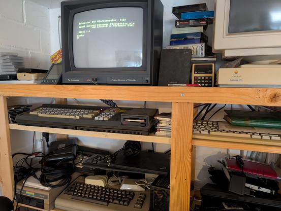 Ein Holzregal in welchem etliche alte Computer stehen. Ein Schneider CPC664 ist eingeschaltet und zeigt auf dem angeschlossenen Monitor CTM644 die Startmeldung. Im Regalfach darunter steht neben einer Commodore Floppy 1541 ein Commodore C64. Darauf liegt einen NoName-Funktastatur und darauf ein IBM ThinkPad. Links daneben liegt eine anthrazitfarbene Commodore Datasette auf einem Commodore +4. Neben dem Schneider Monitor ist ein Amstrad PDA600 und ein Commodore Minuteman Taschenrechner zu sehen. Links neben dem Schneider Monitor steht ein Palm Pilot in einem Dock, neben dem ein Nintendo Gameboy liegt. Im rechten Teil des Fotos ist ein Macintosh Performa 475 mit dazugehörigem Monitor und Tastatur zu sehen. Das Ensemble wird vervollständigt von einer GEOS Mouse aus 1984 und einer IBM Mouse Modell 6450350.