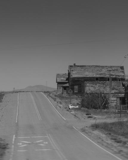 A black-and-white image of a two lane highway with two old wooden houses on the right side. The houses show signs of deterioration, and the landscape is open with distant hills visible in the background.