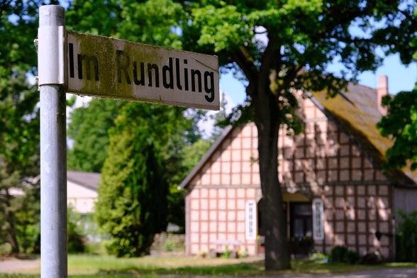 Foto von einem Straßenschild "Im Rundling", dahinter ein großer Baum und ein Fachwerkhaus