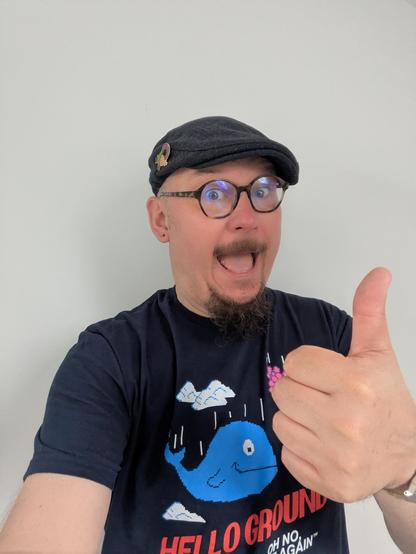 A smiling bespectacled man with a blue peaked cap smiles at the camera, and give a thumbs up. He's got a lilac pin in his hat, and a t-shirt that looks like an 8bit drawing of a smiling whale as it falls through the atmosphere. There's also a bunch of flowers in a pot, thinking "Oh no, not again." 

If we knew why the flowers said that, we'd have a much better understanding of the Universe.
