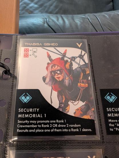 Closeup of a game card in a card holder in a ring binder.
The card shows a cyberpunk'y woman named Thabisa Gbheo. Part of the card is covered by the card holder, that says:

Security Memorial 1
Security may promote one Rank 1 Crewmember to Rank 2 OR draw 2 random Recruits and place one of them into a Rank 1 sleeve.