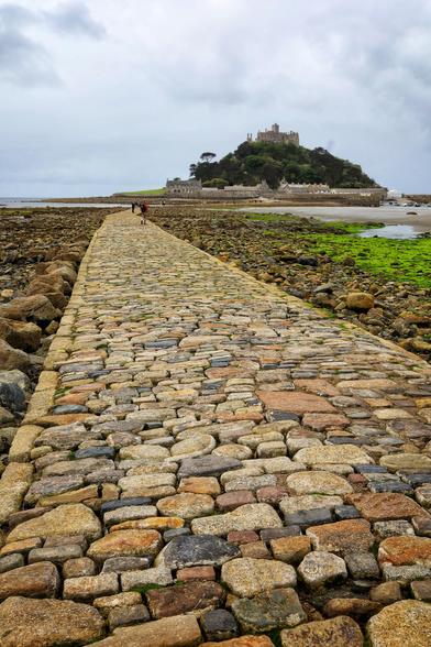 A cobblestone causeway running between seaweed covered stones. At the end of the causeway a castle stands on the top of an island rising high out of the sea.