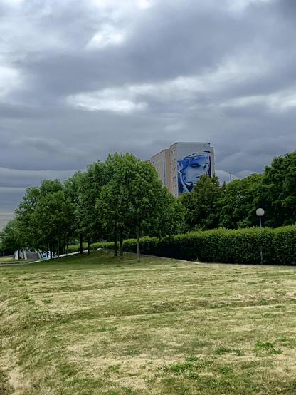Eine Wiese mit ein paar grünen Bäumen. Im Hintergrund stehen ein paar Wohnhäuser. Auf der Fassade des ersten Hauses prangt eine Fassadenmalerei des Kosmonauten Juri Gagarin.