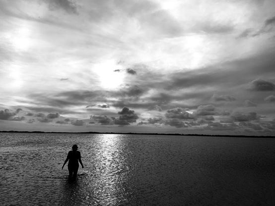 Black and White photo Ocean dark, sunset Sky the sun hidden behind clouds of many hues. A single person silhouetted in the water