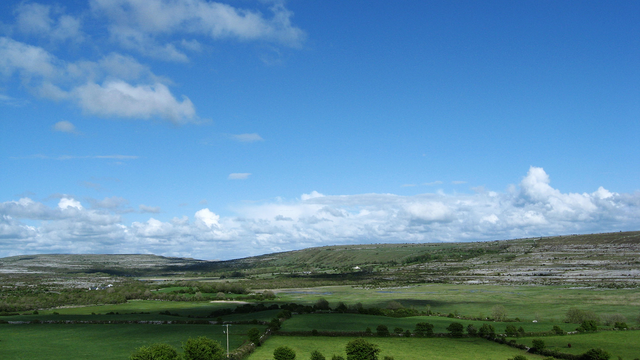 A landscape photograph showing a slightly cloudy blue sky above karst limestone hills with Carron Polje in the foreground, with a patchwork of green fields, hedgerows and trees. A polje,  also called karst polje or karst field, is a large flat depression found in karstic regions. This polje often floods in large parts after heavy rainfall when the groundwater level in the karstic aquifer rises above a certain level. It then looks like a large lake.