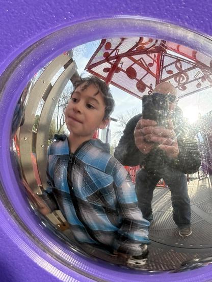 A young child in a plaid jacket is reflected in a circular, shiny surface, along with the silhouette of an adult holding a camera. The background shows a playground structure with musical notes. The scene is outdoors on a bright day.