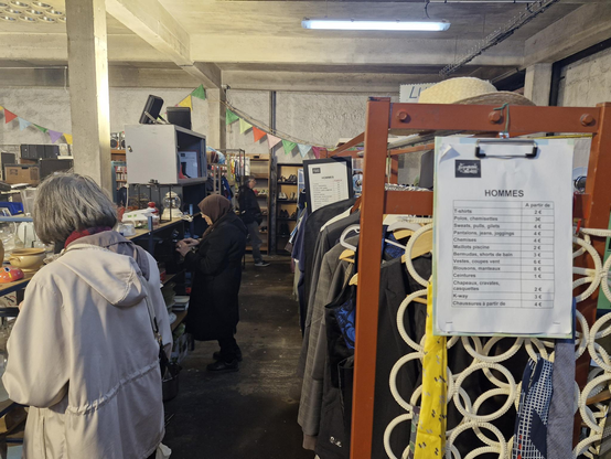 Photo de la Ressourcerie de Malakoff le samedi 24 mai 2025 : un rayon de vestes et de gilets sur la droite, un rayon de vaiselle et d'objets de cuisine sur la gauche, avec deux personnes en train de chiner. 

Sur le rayonnage de droite, on voit les prix affichés des différents vêtements, entre 2€ et 4€.