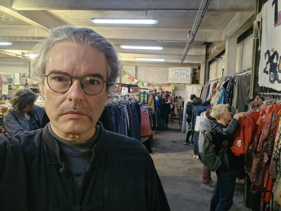 Je pose devant un des rayonnages de la Ressourcerie, l'air bien trop sérieux, avec un gilet noir et les cheveux tirés en arrière et une grosse moustache (pour de vrai). Derrière moi, on distingue plusieurs clients en train d'examiner des vêtements. Tout au fond, on distingue un panneau fixé au mur indiquant "Luminaires, meubles, sport, puériculture".