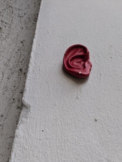 An einer gräulich-weiß verputzten Außenwand ist ein dunkelrotes, künstliches menschliches Ohr angebracht.