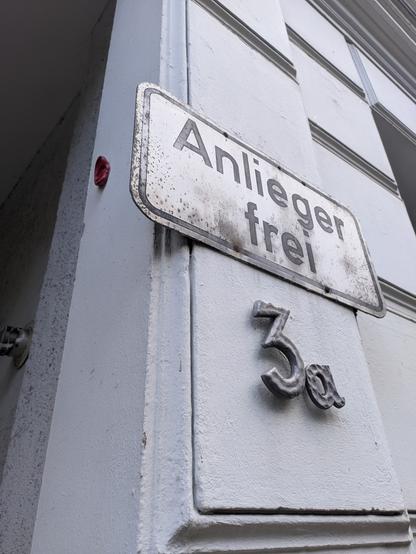In der rechten Bildhälfte ist an der Seite einer Einfahrt schräg ein leicht verwittertes Schild "Anlieger frei" zu sehen, darunter die Hausnummer "3a". In der linken Bildhälfte ist zu Beginn der Einfahrt klein ein dunkelrotes künstliches menschliches Ohr neben bzw. hinter dem Schild "Anlieger frei" zu sehen.