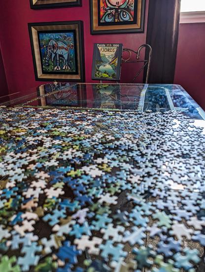 We're at a low angle looking diagonally along a glass dining table on which the edge pieces of a jigsaw have been put together at the far end while the end near us has all the remaining puzzle pieces face up and laid out randomly ready for fitting. The dominant colours of the jigsaw are blues, teals, and greens. The glass table reflects light from a window in the wall behind and high up and it shows that there are lots of fingerprints on the glass surface at the end. The window has long, dark brown curtains hanging down, and the wall is painted a deep raspberry. A few abstract, vibrant, cartoon or street art-style pieces of artwork are mounted on the wall and an elephant figure is easy to make out. These pieces are also reflected in the glass at the end of the table. A metal-framed chair is at the far end and the jigsaw box is hanging from the backrest, facing us. It shows the puzzle is that of a cruise ship in Norwegian fjords in a style that would not be too out of place in 1930s art deco travel posters.