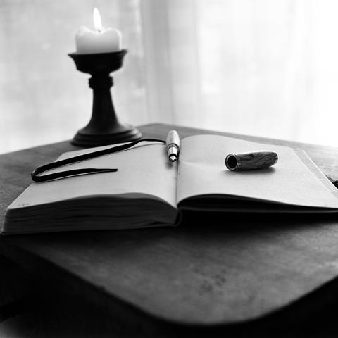 A black and white still life photograph captures a lit candle, an open notebook, and a pen on a dark, possibly wooden, table. The candle, burning brightly, sits in a dark, ornate candle holder on the left side of the frame. To its right, an open notebook lies flat, with its pages facing upwards. A pen with a metallic tip and a darker, textured body rests on the left page of the notebook, its cap detached and placed on the right page. A dark ribbon or bookmark extends from the left side of the notebook. The background is softly lit, suggesting a window or light source, creating a gentle glow behind the objects. The overall mood is contemplative and classic.