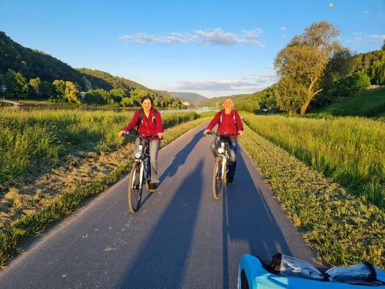 2 Menschen sitzen auf dem Fahrrad und fahren auf dem Elberadweg in der sächsischen Schweiz an der Elbe der Abendsonne entgegen.