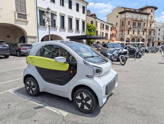 Photo of an XEV Yoyo microcar parked in an motor scooter parking in an Italian piazza.