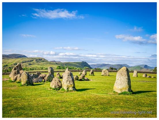 Part of a circle of various sized upright boulders in an area of short grass, lit by low angle sunlight, with a mainly clear blue sky.