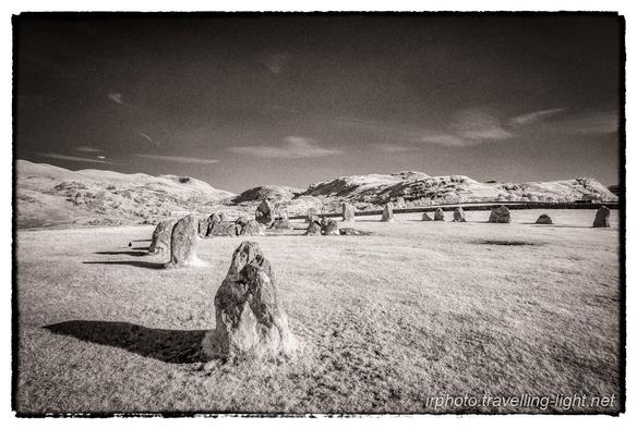 A toned black and white infrared photo showing part of a circle of various sized upright boulders in an area of short grass, lit by low angle sunlight, with a mainly clear blue sky.