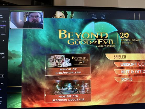 Das Bild zeigt den Startbildschirm des Spiels "Beyond Good & Evil 20th Anniversary Edition" auf einem Computermonitor. Der Hintergrund ist ein kosmischer Hintergrund mit einem großen, glänzenden Planeten und einem Farbverlauf von Grün zu Rot. Im Zentrum des Bildschirms ist der Titel "Beyond Good & Evil" in goldenen Lettern zu sehen, gefolgt von "20th Anniversary Edition" und "A Ubisoft Original".
Auf der linken Seite des Bildschirms befinden sich zwei Optionen: "Besuche die Jubiläumsgalerie" mit einem Bild, das verschiedene Illustrationen und Fotos zeigt, und "Probier den Speedrun-Modus aus" mit einem Bild, das eine Szene aus dem Spiel zeigt. Rechts auf dem Bildschirm sind die Optionen "SPIELEN", "UBISOFT.COM", "HILFE & OPTIONEN" und "BONUS" in orangefarbenen Rechtecken angezeigt.
Im oberen linken Eck des Bildschirms ist ein kleines Fenster mit einem Mann zu sehen, der ein Kopfhörer trägt und in die Kamera schaut. Der Bildschirm zeigt auch verschiedene technische Informationen wie "90/140" und "66/140" in der linken oberen Ecke.