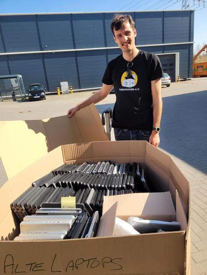 Junge Person vor einem Karton mit sehr vielen Laptops. Die männlich gelesene Person trägt ein T-Shirt mit Aufdruck "Computertruhe e.V.", auf dem Karton steht "Alte Laptops".