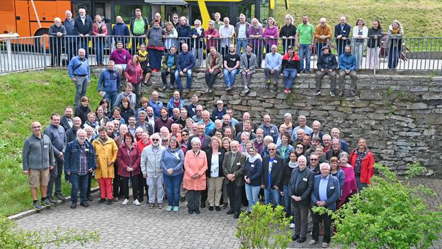 Das Bild zeigt die Konferenzmitglieder, die für ein Gruppenfoto posieren. Sie stehen im Freien vor einer niedrigen Steinmauer. Im Hintergrund ist ein gelber Reisebus zu sehen. Die Menschen sind unterschiedlich alt und tragen verschiedene Kleidung.