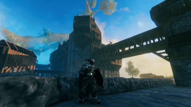 Valheim Screenshot: Ein Wikinder steht mit Schwert und Schild an einer hüfthohen Mauer. Dahinter ist ein großes Gebäude zu sehen, zu dem eine hohe, überdachte Brücke führt.