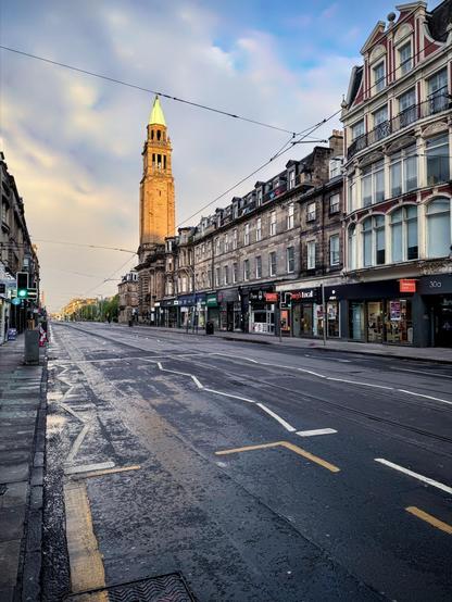 This image shows a city street scene, likely early in the morning or late in the afternoon given the lighting. The wet streets and muted light suggest recent rainfall or an early morning scene before the city has fully awakened. The architecture is distinctly European, with historic building facades and a prominent stone tower that stands out in the skyline, possibly indicating a church or old municipal building.

The street is lined with various shops and has tram lines embedded into the road, indicating a city with a public transit system. The absence of people and cars lends a serene, almost deserted atmosphere to the scene. The image captures a blend of historic charm and modern urban life.